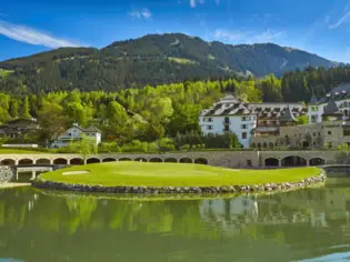 Ein idyllischer Golfplatz mit einem gepflegten Grün, umgeben von einem Teich und einer beeindruckenden Berglandschaft. Im Hintergrund steht ein elegantes Hotelensemble mit traditioneller Architektur, eingebettet in die Natur.