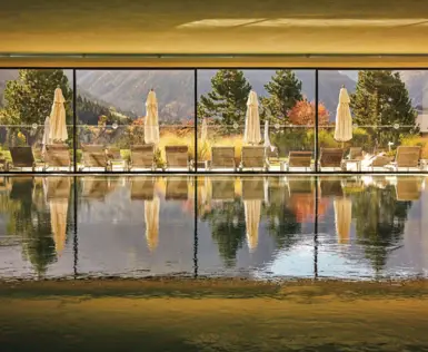 Lichtdurchfluteter Innenpool im A-ROSA Ifen mit Blick auf Liegestühle, Sonnenschirme und die herbstliche Berglandschaft des Kleinwalsertals durch große Panoramafenster.
