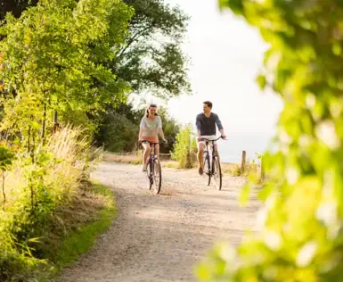 Zwei Personen fahren auf einem Schotterweg Fahrrad, umgeben von Bäumen.
