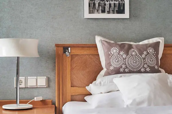 A modern designed hotel room with a focused view of a bed with a wooden headboard and a decorative pillow with a delicate floral pattern in front of a light gray wallpapered wall . On the bedside table next to the bed is a stylish table lamp with a white lampshade next to a row of sockets and light switches on the wall. A framed black and white photograph hangs above the bed, adding to the simple and elegant interior of the room.