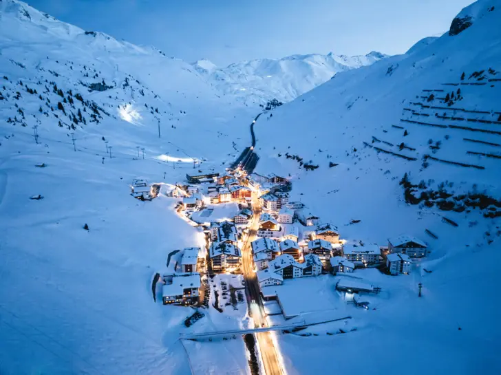 Verschneites Bergdorf bei Dämmerung, umgeben von hohen schneebedeckten Bergen. Die beleuchteten Straßen und Häuser des Dorfes erzeugen einen warmen Kontrast zur kalten, blauen Winterlandschaft.