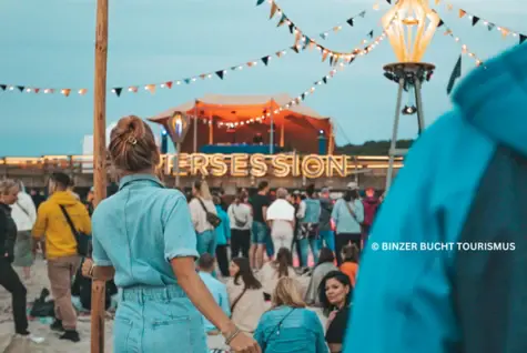 Auf einem Pier findet ein Fest statt. Bunte Girlanden mit kleinen Lichtern sind über den Platz gespannt und viele Menschen tummeln sich. 