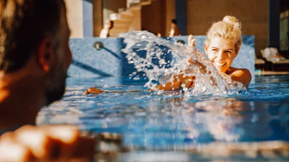 Eine Frau planscht lächelnd Wasser in Richtung eines Mannes. Beide schwimmen im Pool, im Hintergrund sind weitere Personen auf Liegen. 