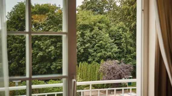 Blick durch ein Fenster auf Bäume und ein Gebäude bei Tageslicht.