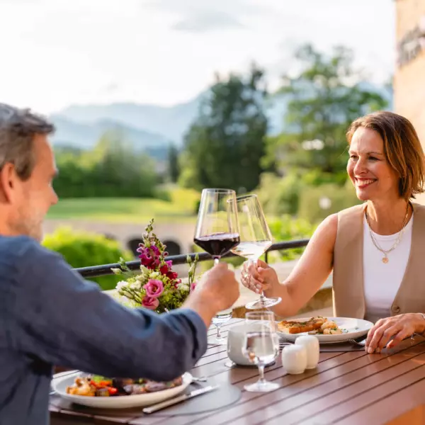ALT-Text: "Ein Paar genießt ein romantisches Abendessen auf einer Terrasse mit idyllischem Bergblick. Sie stoßen mit Weingläsern an, während sie von einer entspannten Atmosphäre umgeben sind, die durch die warmen Sonnenstrahlen der Abenddämmerung und das satte Grün der umliegenden Landschaft verstärkt wird.