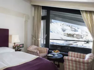 A hotel room with a bed and two armchairs and a large window with a view of a snow-covered mountain landscape.