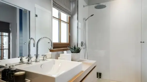 Ein modernes Badezimmer mit einem eckigen Waschbecken auf einem hölzernen Waschtisch und im Hintergrund ist eine verglaste Duschkabine zu sehen. 