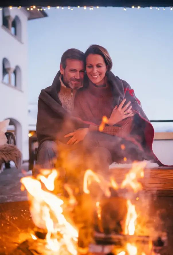 Ein Paar sitzt lächelnd Arm in Arm unter einer Decke an einem kleinen Feuer.