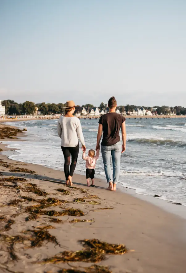 Zwei Eltern halten ihr kleines Kind zwischen sich an den Händen und spazieren am Strand entlang. Das Ambiente ist in warme, ruhige Farben getaucht. 