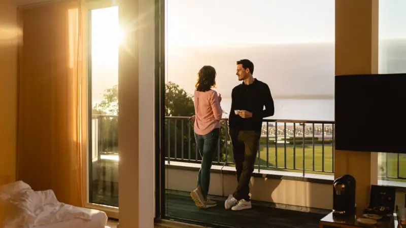 Ein Mann und eine Frau stehen auf einem Balkon und blicken aus einem Fenster.