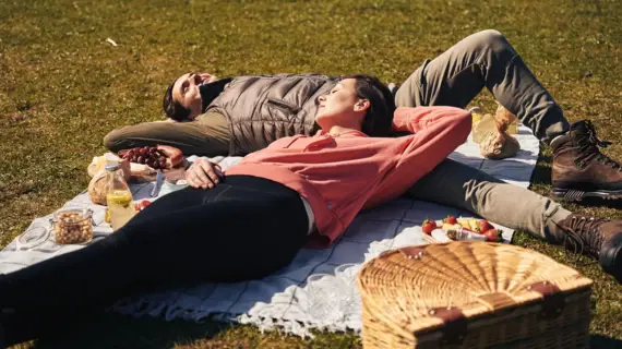Paar liegt entspannt auf einer Picknickdecke im Grünen, umgeben von Snacks, Getränken und einem Picknickkorb