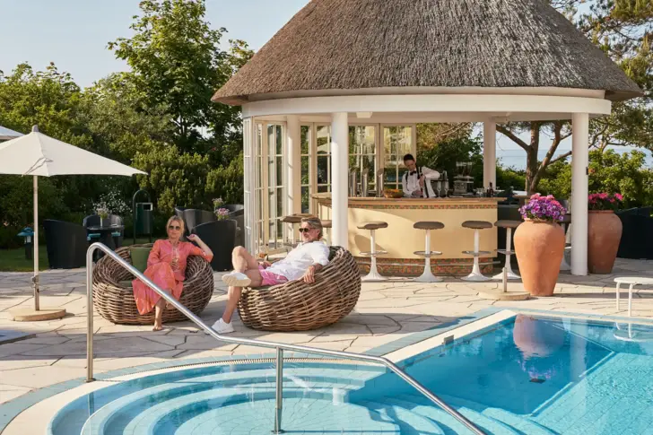 Ein Mann und eine Frau sitzen in zwei Sesseln an einem Außenpool mit Poolbar im Hintergrund. 