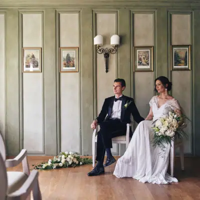 Ein Mann und eine Frau sitzen in Stühlen, die Frau trägt ein Brautkleid und hält einen Blumenstrauß.