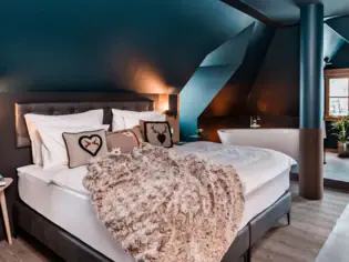 Ein gemütliches Bett mit drei Dekokissen im alpenstil steht an einer dunkelblauen Wand. Es liegt eine braune Kuscheldecke vorne auf dem Bett und im Hintergrund ist eine freistehende Badewanne zu sehen. 