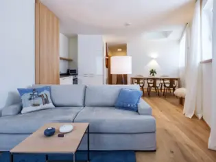 Ein Wohnbereich mit einem grauen Sofa vor dem ein Couchtisch aus Holz steht. Im Hintergrund ist beine offene Küche sowie ein Esstisch mit sechs Stühlen zu sehen.