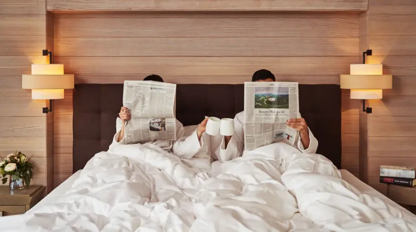 Two people are lying in a bed, reading newspapers and drinking coffee.