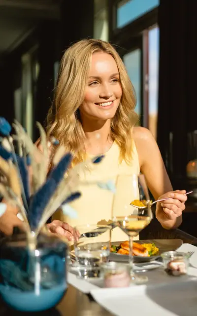 Frau sitzt lächelnd an einem Tisch mit Speisen und einem Weinglas.