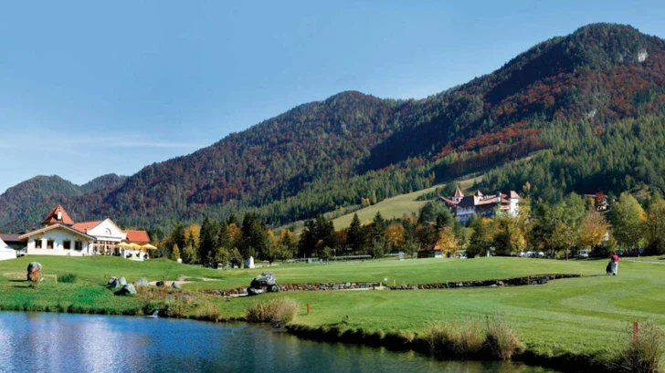 Eine Ansicht der Landschaft mit Teil des großen Teiches, grünen Wiesen, dem Hotel hinter den Bäumen und weiten Bergen. 
