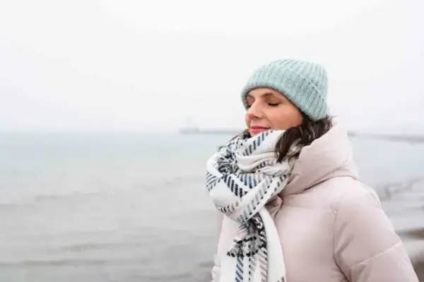 Eine Frau mit Hut und Schal steht am Wasser.