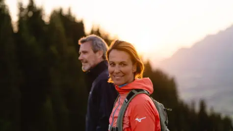 Eine Frau und ein Mann befinden sich in Outdoor-Kleidung auf einer Wanderung in den Bergen. Im Hintergrund geht die Sonne unter.