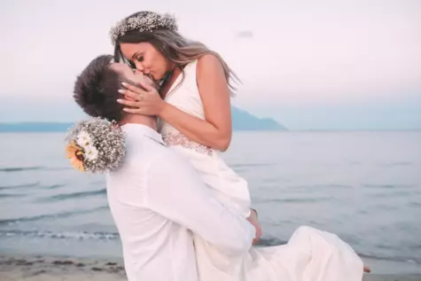 Ein frisch getrauter Bräutigam hebt seine Frau hoch und beide küssen sich am Strand. Das Meer ist ruhig im Hintergrund.