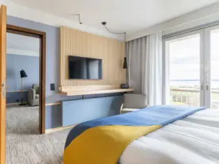 Blick von einem großen Bett mit einer blau-gelben Tagesdecke auf einen Fernseher und in einen Wohnbereich mit Sofa..