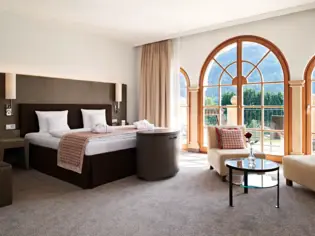 A hotel room with a large bed on the left and a sitting area on the right. Opposite are 3 large, floor-to-ceiling windows with access to a terrace.