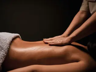 A relaxing wellness massage in a spa setting, where a masseuse's hands gently glide over the back of a woman lying on a massage table. A soft towel covers the lower part of the woman while her skin glistens from the massage oil. The dark background emphasizes the peaceful and calming atmosphere of the scene.
