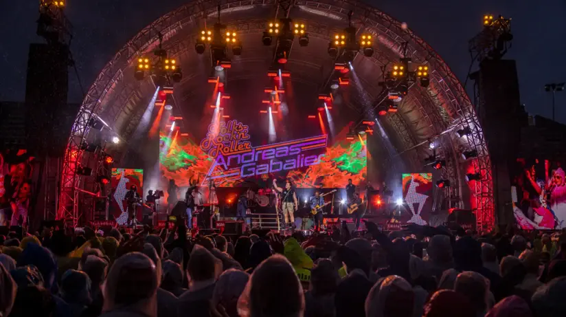 Ein lebhaftes Open-Air-Konzert bei Nacht mit einer Band, umgeben von dynamischen Lichtern und einer Menschenmenge, die zuschaut. 