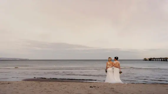 Ein Brautpaar steht mit umgelegten Armen in ihren weißen Kleidern und ihrem Brautstrauß in der Hand am Strand und blicken auf das ruhige Wasser. 