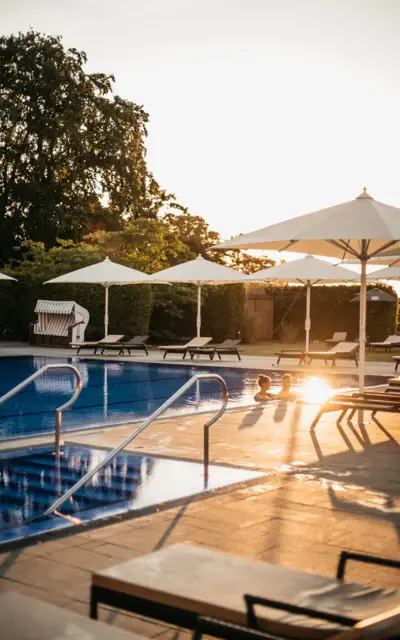 Ein Poolbereich im Freien bei Sonnenuntergang mit aufgespannten weißen Sonnenschirmen, Liegestühlen und sichtbaren Pooltreppen, wo das Licht der untergehenden Sonne sich im Wasser spiegelt und zwei Personen im Pool schwimmen.