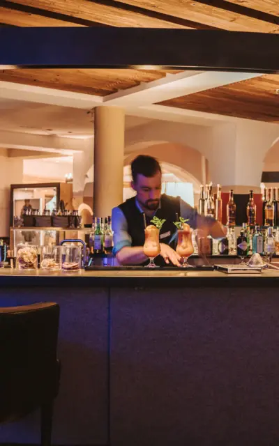 Ein Barkeeper in einem stilvollen, gemütlich beleuchteten Barbereich bereitet einen Cocktail zu. Der Tresen ist mit verschiedenen Flaschen und Gläsern bestückt. Im Hintergrund sind weitere Gäste und die elegante Einrichtung der Bar zu sehen.