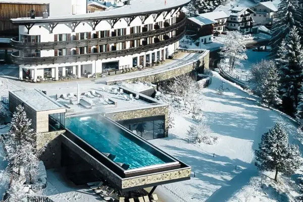 Building with a pool in the middle of a mountain village, surrounded by trees and a landscape with mountains.