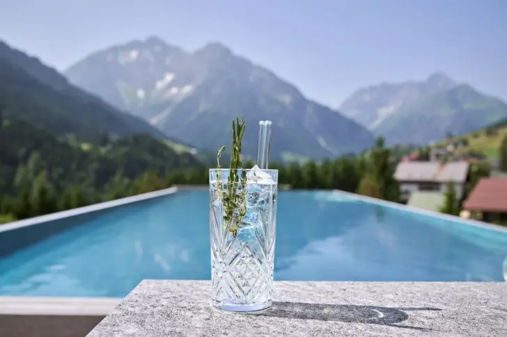 An dem Beckenrand eines Pools steht ein Gin Tonic. Die Sonne scheint warm auf das Wasser und im Hintergrund sind Berge zu sehen. 