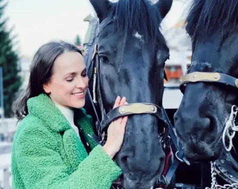 A woman in a green coat strokes two horses with harnesses.