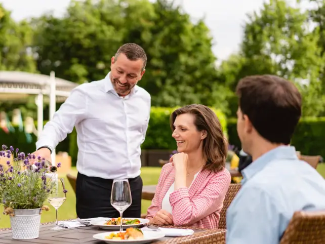 Ein Mann und eine Frau sitzen an einem Tisch im Grünen. Vor ihnen stehen zwei Teller befüllt mit Speisen, zwei Weingläser und ein Blumentopf mit Lavendel auf dem Tisch. Ein Kellner in einem weißen Hemd steht neben der Frau und gießt Wein in ihr Glas.