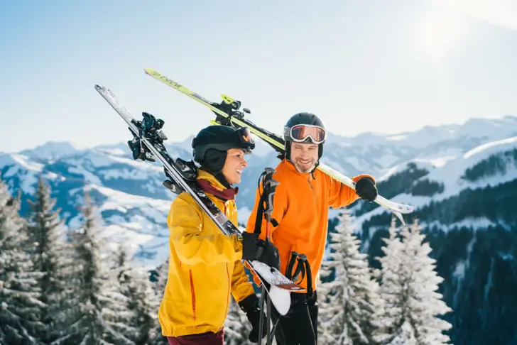 Zwei Personen in Skikleidung mit Skiern auf einem verschneiten Berg mit schneebedeckten Tannen im Hintergrund.