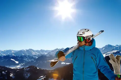 Mann mit Skihelm und -brille hält Skier auf der Schulter auf dem Gipfel vor einer verschneiten Bergkulisse und blauem Himmel mit strahlender Sonne.