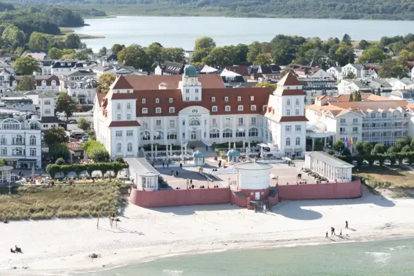 Großes weißes Gebäude im Stil der Bäderarchitektur neben einem Gewässer mit Bäumen und Booten.