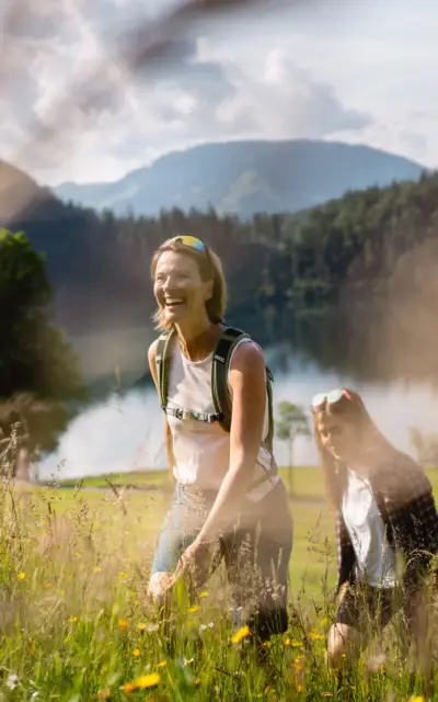 Zwei Frauen wandern in einem Feld, umgeben von Gras und Blumen vor einer Bergkulisse.