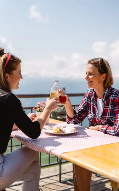 Zwei Frauen sitzen an einem Tisch im Freien und stoßen mit Getränken an.