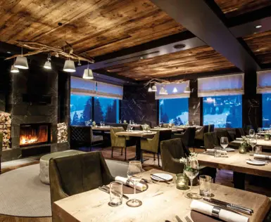 Ein stilvoll eingerichtetes Restaurant mit rustikalen Holzelementen, gemütlichen grünen Sesseln und elegant gedeckten Holztischen. Ein Kamin mit brennendem Feuer sorgt für eine warme Atmosphäre, während große Fenster einen beeindruckenden Blick auf eine schneebedeckte Berglandschaft bieten.