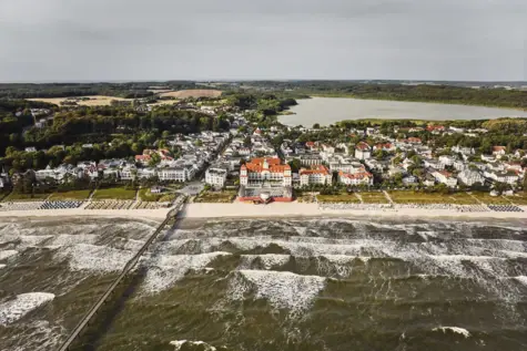 Das Seebade Binz aus der Luft fotografiert. Im Vordergrund sind die Wellen und das Meer, dahinter befindet sich Binz mit viel hellem Gebäuden und ein großer See. 