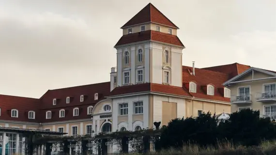 Eine Außenansicht des Kurhauses Binz bei Sonnenuntergang. Die hellen Fassenden und das rote Dach sind von der Sonne in warme Farben getaucht und der Himmel ist hell bewölkt. 