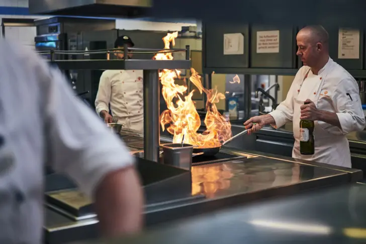 In einer Restaurantküche stehen drei Köche in weißen Kochschürzen und bereiten Speisen zu. Einer von ihnen tut dies mit hohen Flammen in seiner Pfanne und einer Flasche in seiner Hand. 