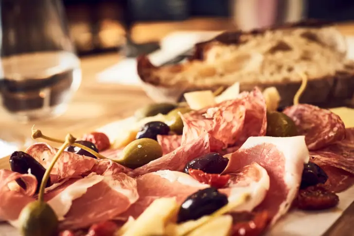 Ein Holzbrett mit einer Variation aus Oliven, Käse, Salami und getrockneten Tomaten. Daneben steht zwei Weißweingläser. 