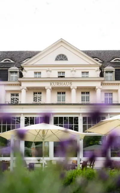 Das weiße Kurhaus in elegantem Stil mit Säulen auf der ersten Etage und Sitzmöglichkeiten aus Holz, für die weiße Sonnenschirme Schatten spenden. 
