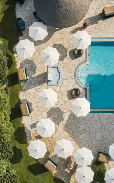 Pool mit Liegestühlen auf der Dachterrasse eines Gebäudes, umgeben von Gras und Pflanzen, aufgenommen aus der Vogelperspektive.