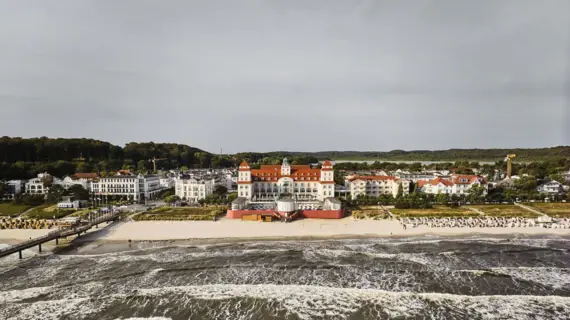 Eine Vogelperspektive des Kurhauses Binz und dem Seebad mit vielen weißen Gebäuden und dem rauschenden Meer. 