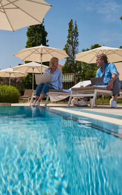 Ein Mann und eine Frau sitzen auf Liegestühlen am Pool unter einem Sonnenschirm.